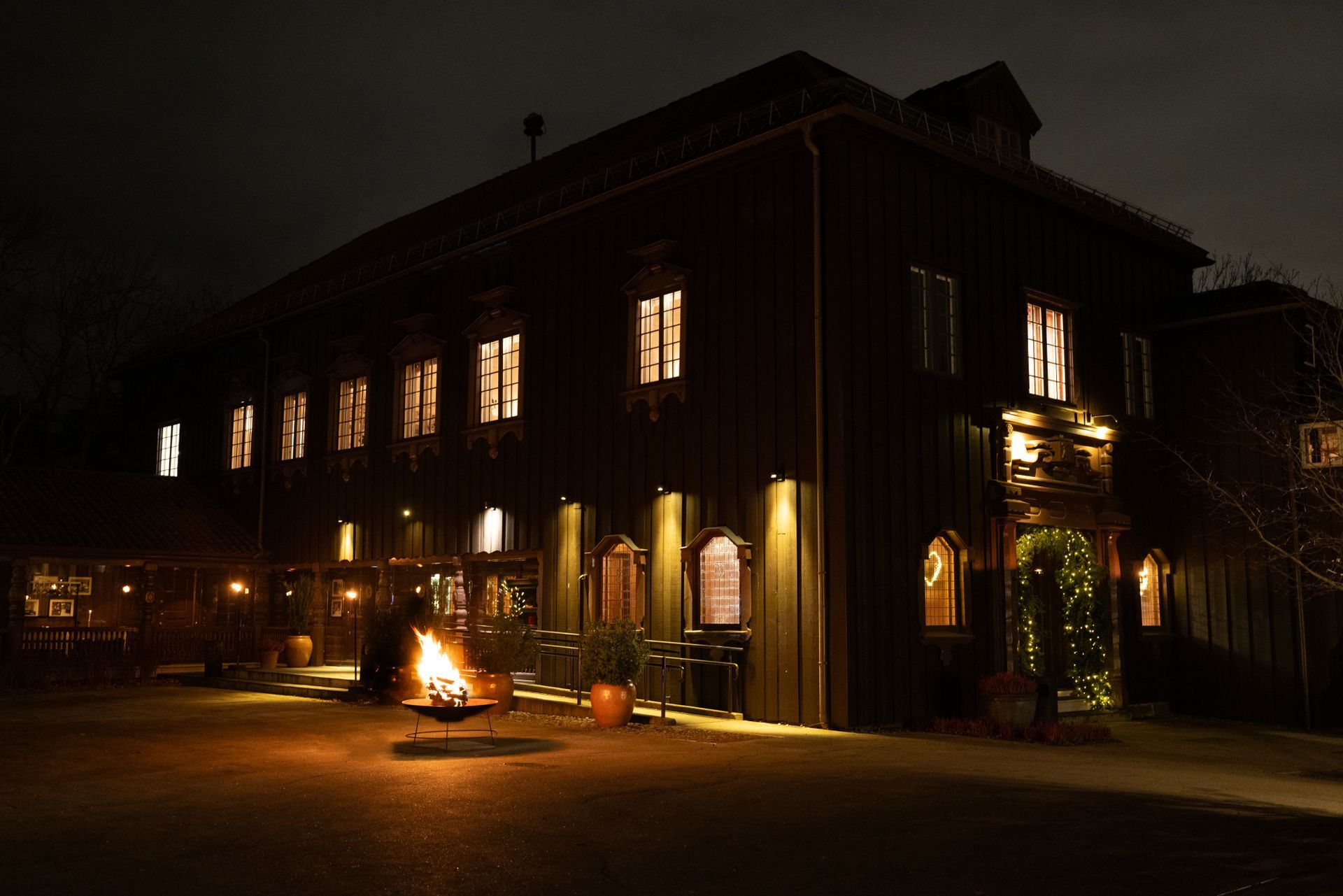 Mørk trebygning om natten med opplyste vinduer og utendørs bålplass.