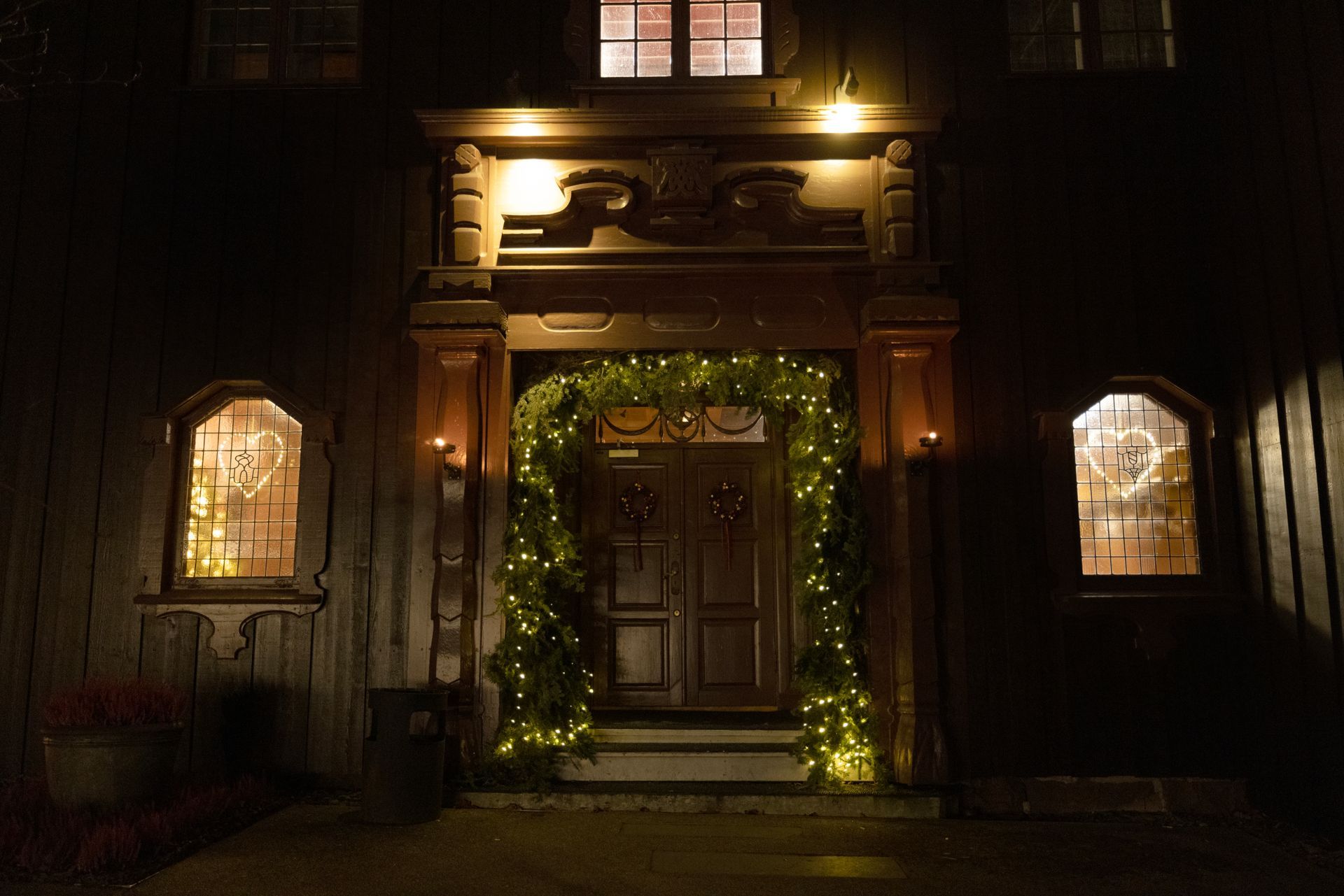 Fasade av trebygning på kveldstid med opplyst døråpning utsmykket med grøntområder og lys.