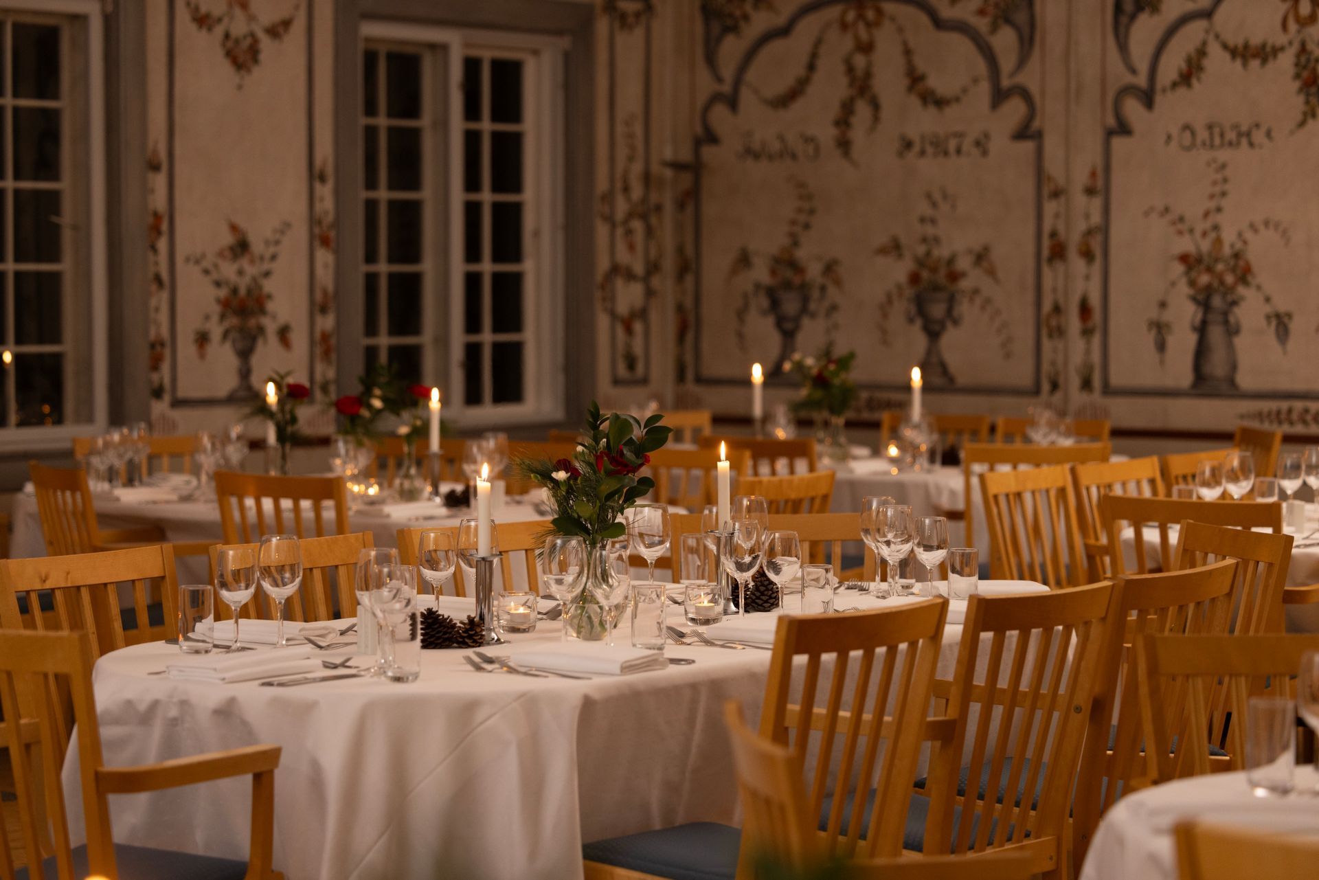 Elegant spisestue, bord dekket med hvit duk, stearinlys og blomsteroppsatser. Lyse trestoler, dekorative veggmalerier.