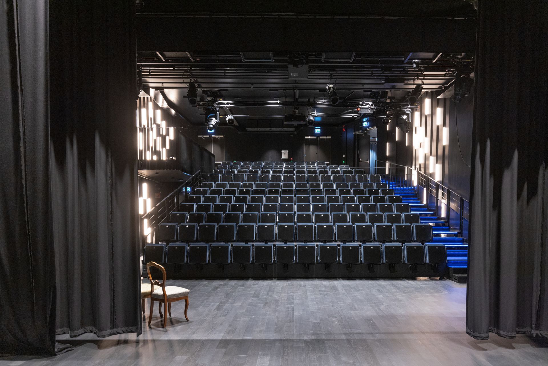 Utsikt over et mørkt teater fra scenen; rader med seter vender mot baksiden. Svarte gardiner rammer inn bildet.