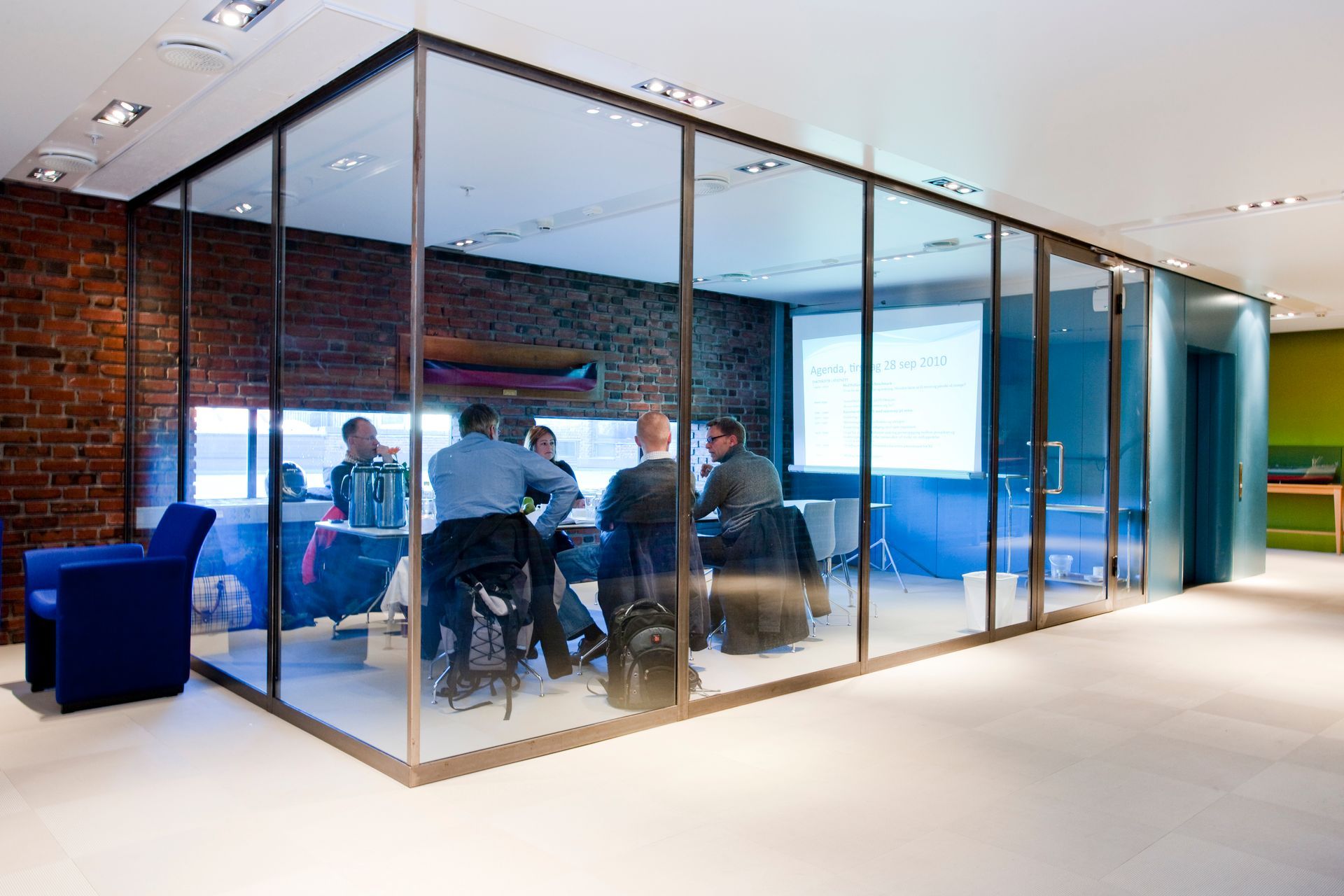 Møterom med glassvegger og folk sittende rundt et bord, presentasjon på skjerm. Murvegg på den ene siden, blå stol utenfor.