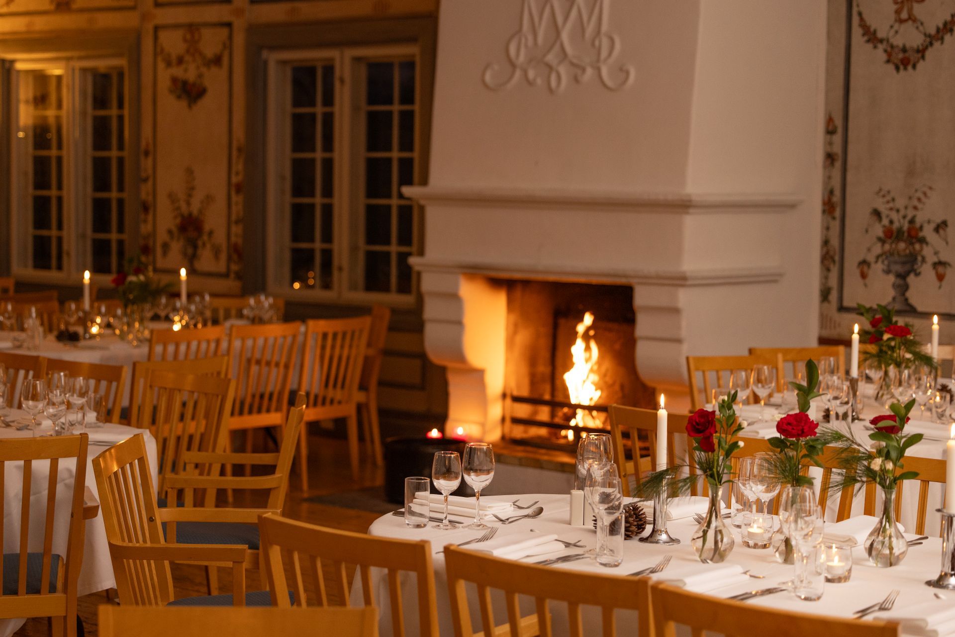 Elegant spisestue med dekkede bord for et måltid, peisbrenning og blomsterdekorasjon på veggene.