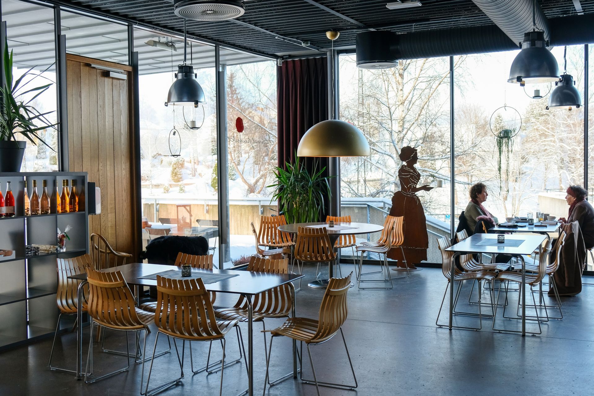 Innvendig utsikt over en kafé med bord, stoler og store vinduer med utsikt over et snødekt utendørslandskap.
