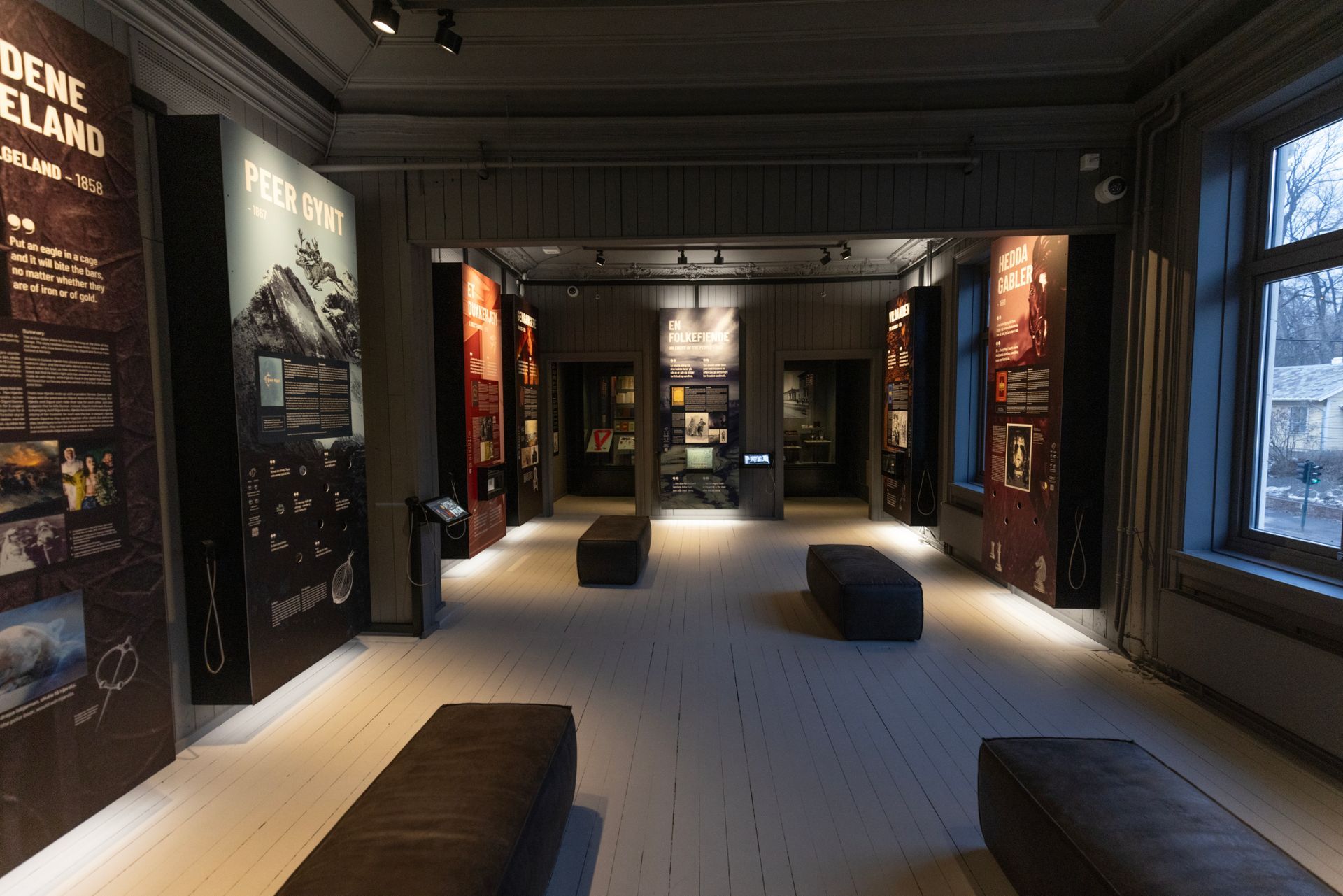 Museumsinteriør med mørke vegger, utstillinger, benker og vinduer.