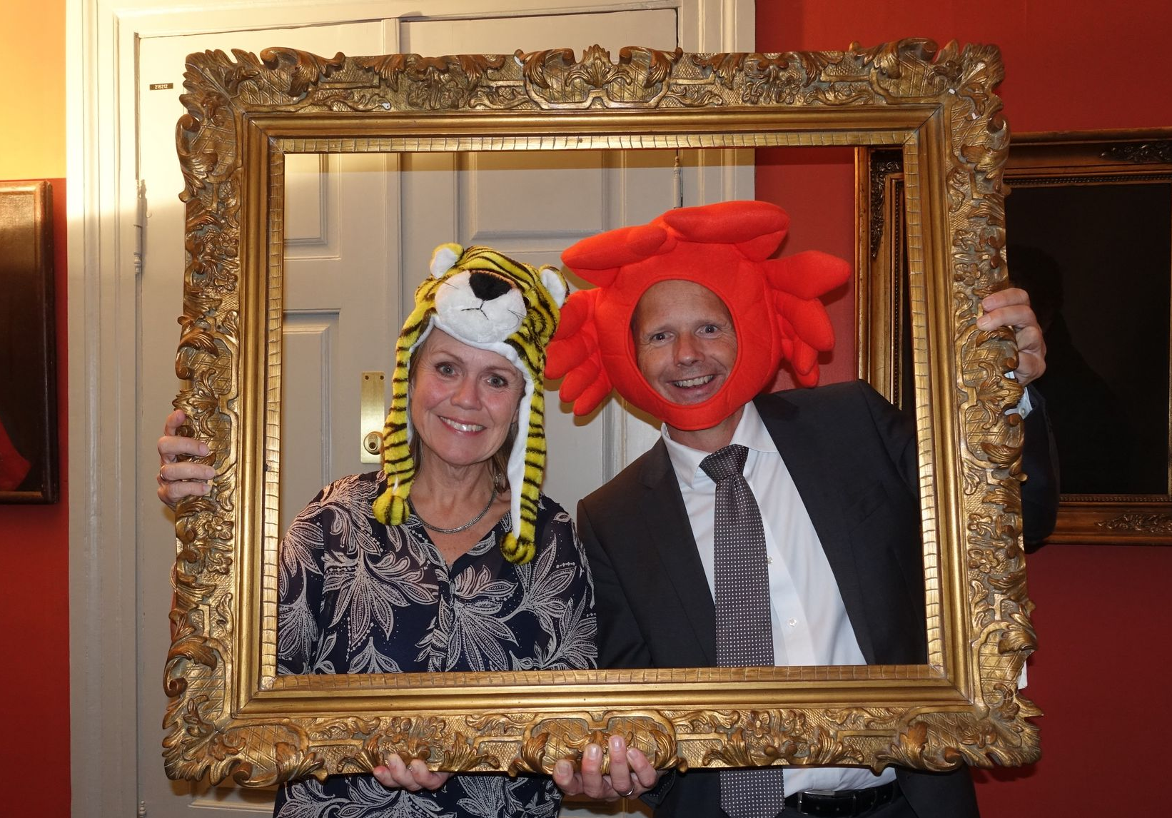 To personer med tullete hatter innrammet av en gullfarget bilderamme, smilende. Rom med røde vegger.