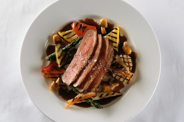 Skiver av biff med grillede grønnsaker i en mørk saus på en hvit tallerken.