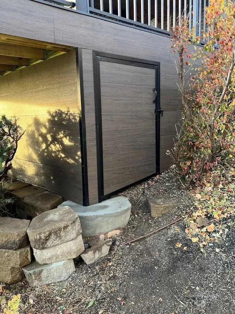 A shed is sitting under a deck next to a rock wall.