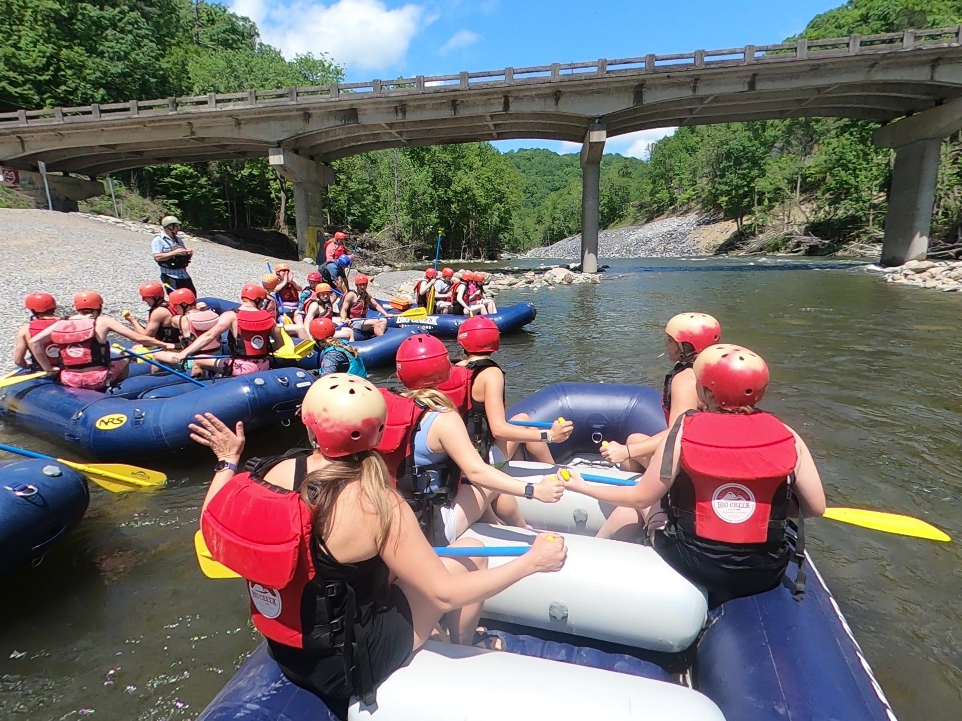 People in red life vests and helmets sit in blue rafts on a river near a bridge on a sunny day.