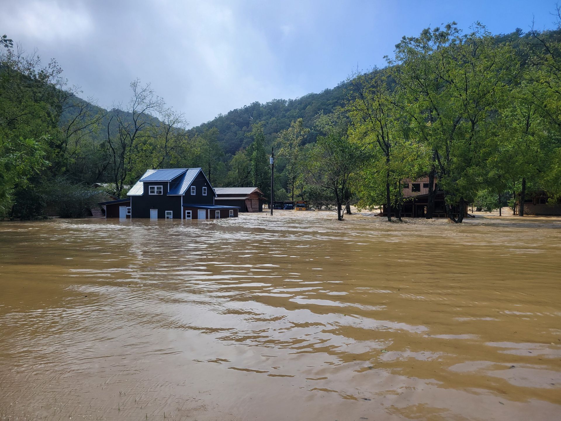 A dark-colored house sits partially submerged in muddy floodwaters, surrounded by trees and a hillside under a cloudy sky.