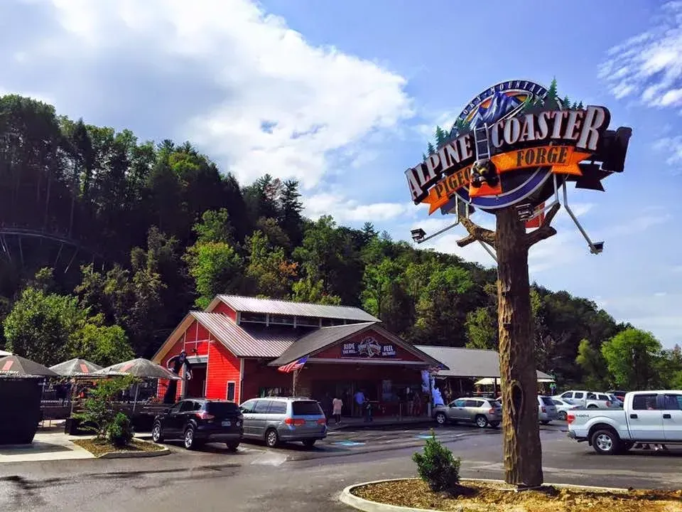 A large sign for Alpine Coaster Pigeon Forge stands in a parking lot in front of a red building and a wooded hillside.