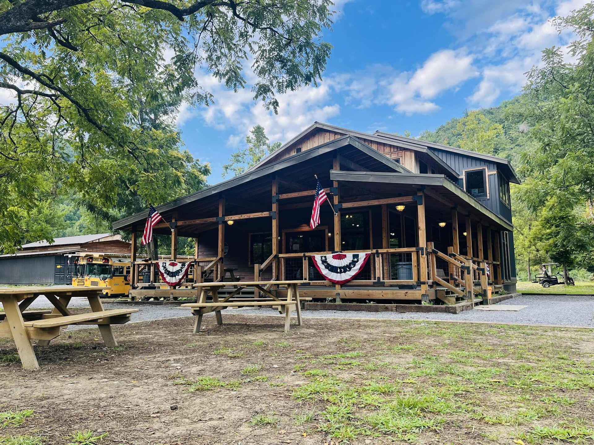 A rustic wooden building with a wrap-around porch decorated with American flags, nestled in a grassy, wooded area.
