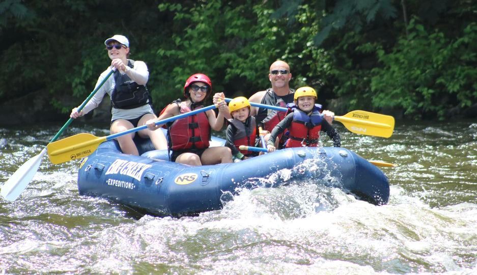 A group of five people in life vests smiling while white-water rafting in a blue raft on a river.