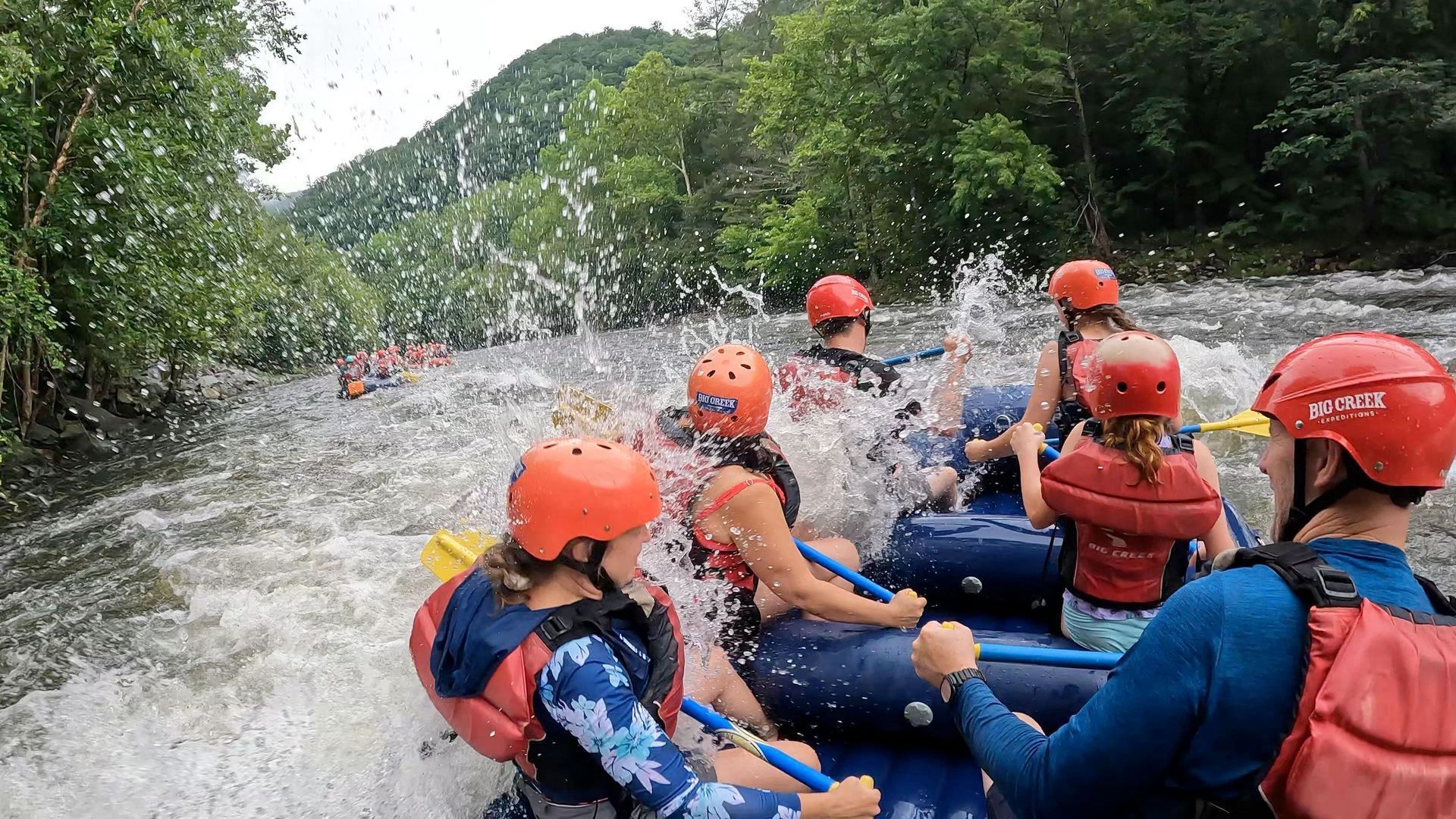 A group of people wearing helmets and life jackets white-water rafting down a river with splashes of water in the air.