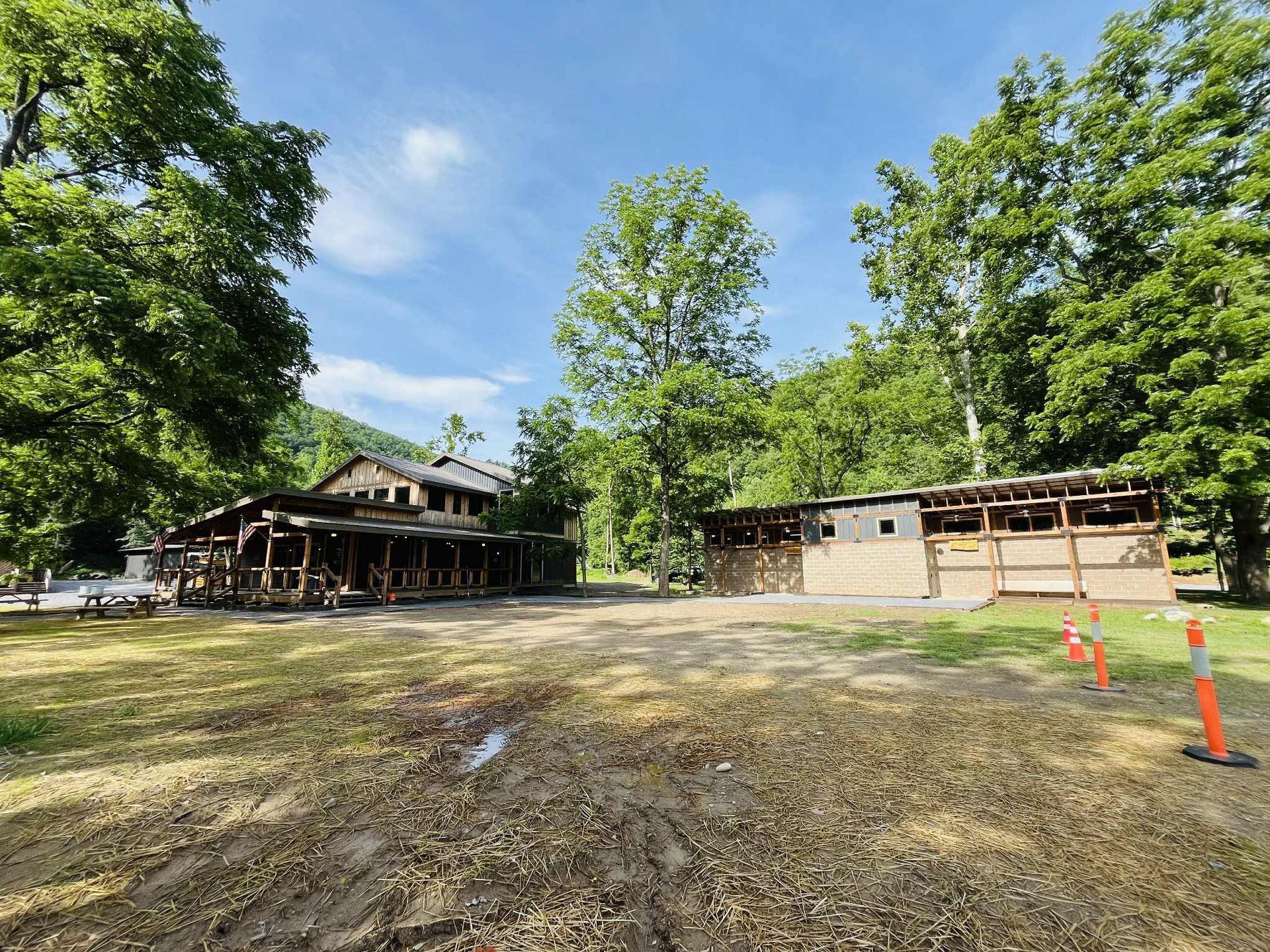 A sunny outdoor scene featuring two partially constructed wooden buildings in a park-like setting with green trees.