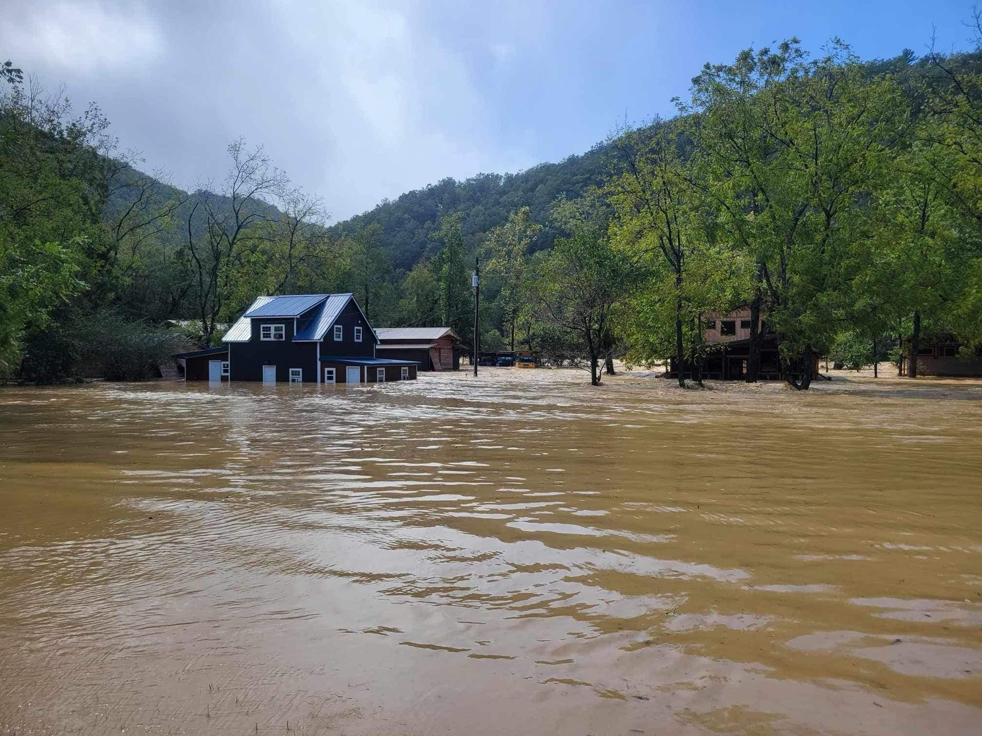 A dark blue house stands partially submerged in deep, muddy floodwaters surrounded by trees and a mountainous backdrop.
