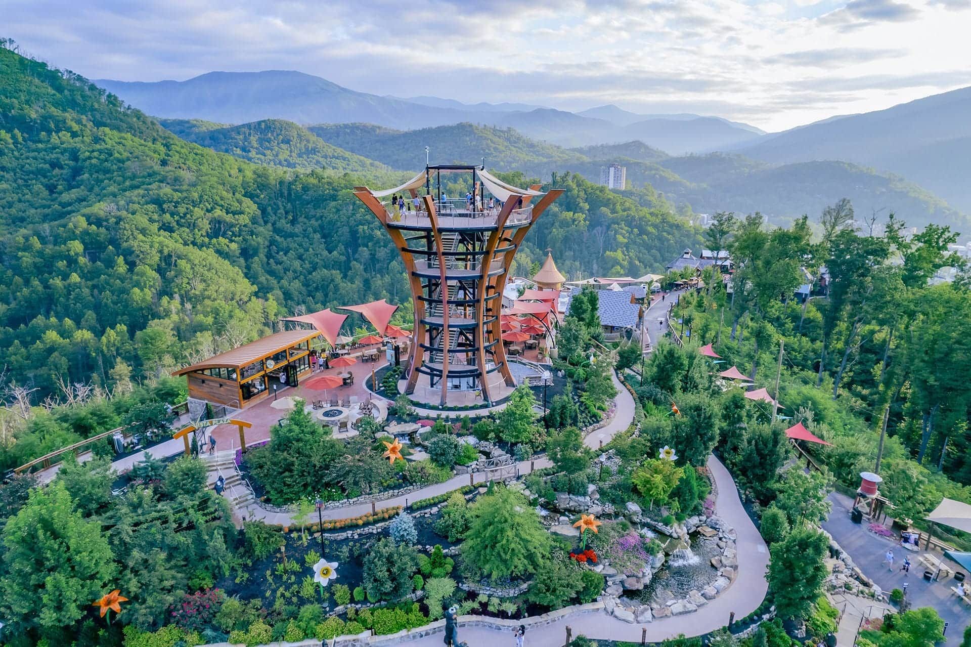 A tall, wooden observation tower stands atop a lush, landscaped hill surrounded by dense forest and rolling mountains.