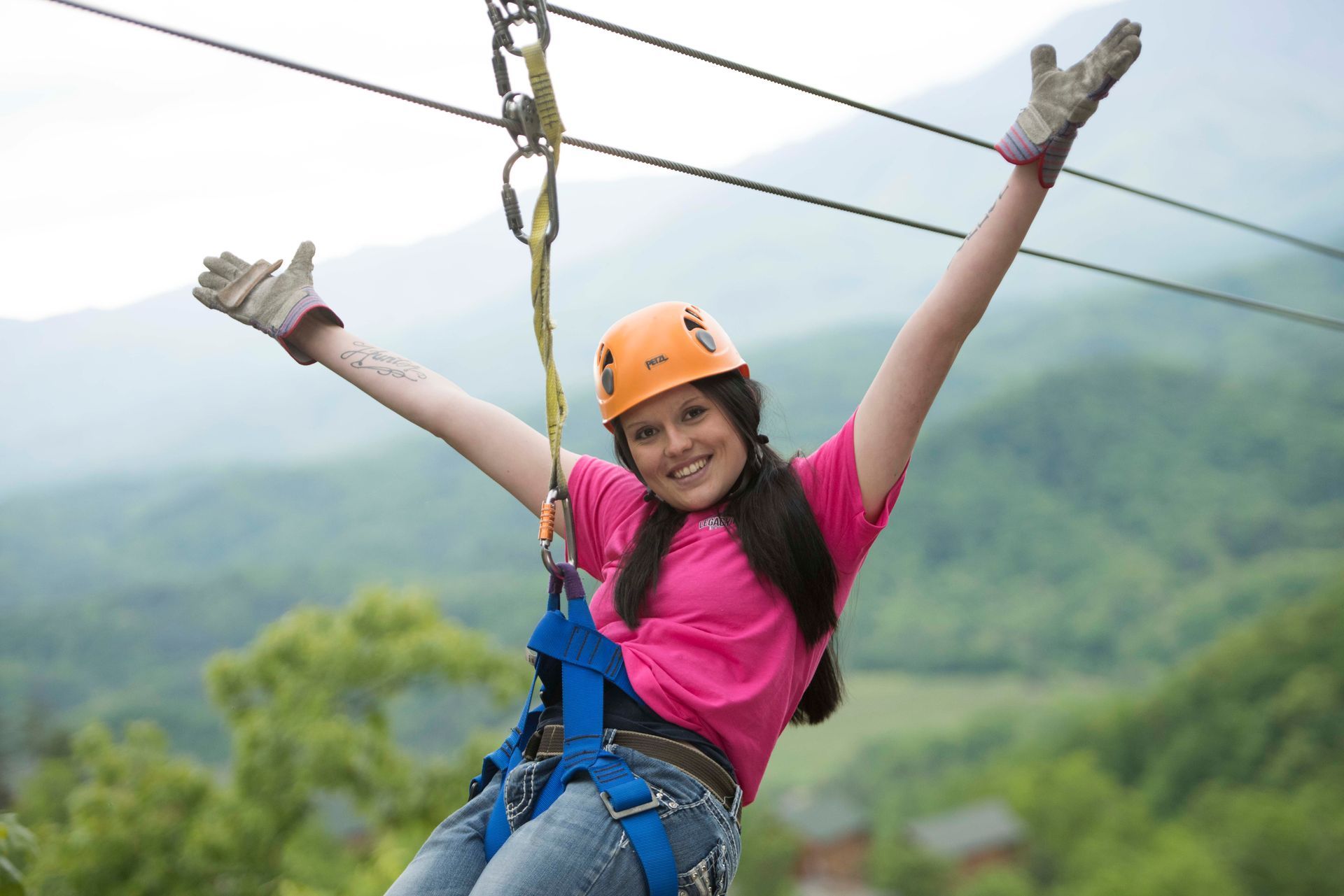 A person in a helmet and pink shirt ziplining with arms outstretched against a backdrop of green mountains.