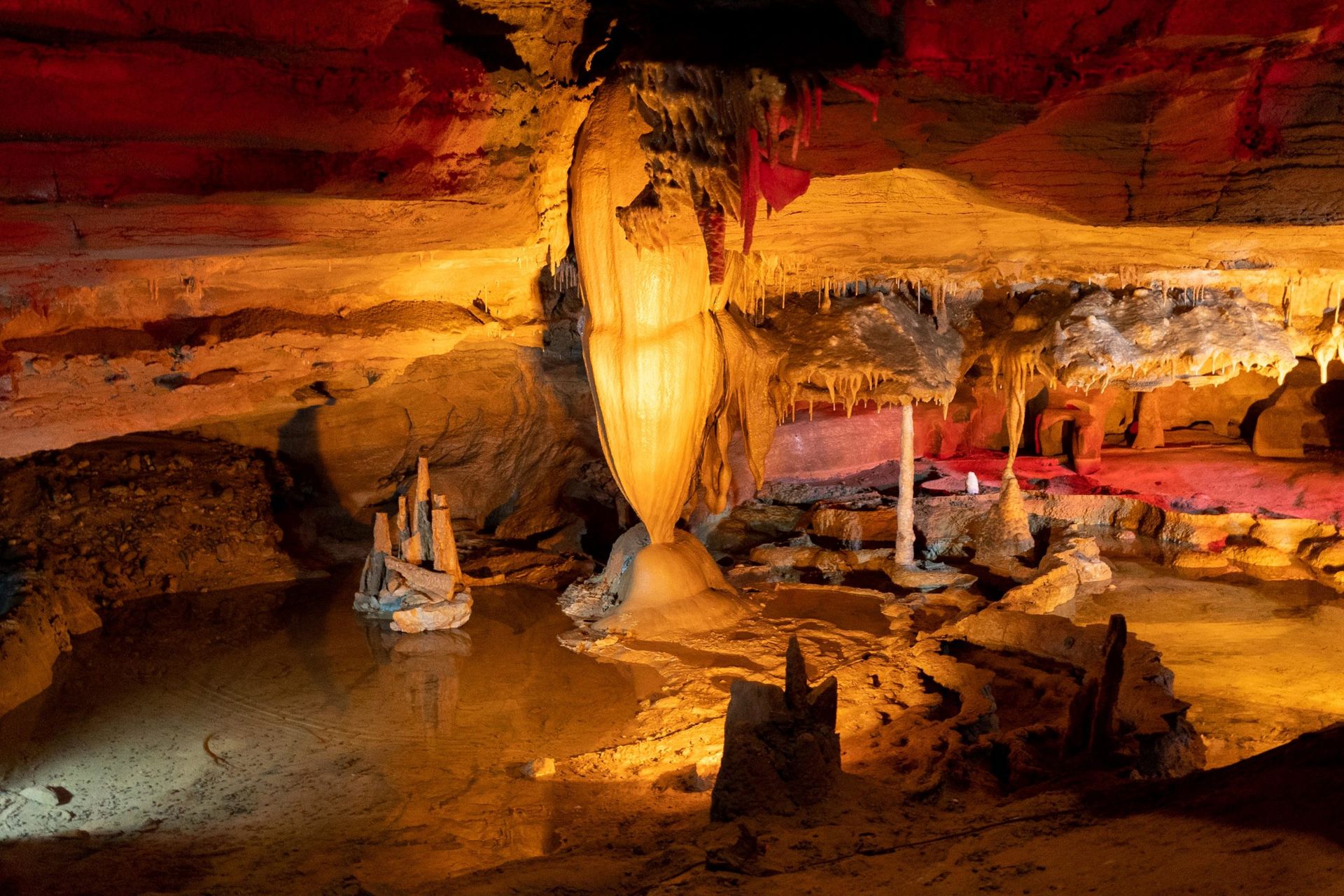 An illuminated limestone cave interior features a large central stalactite formation over a pool of water.