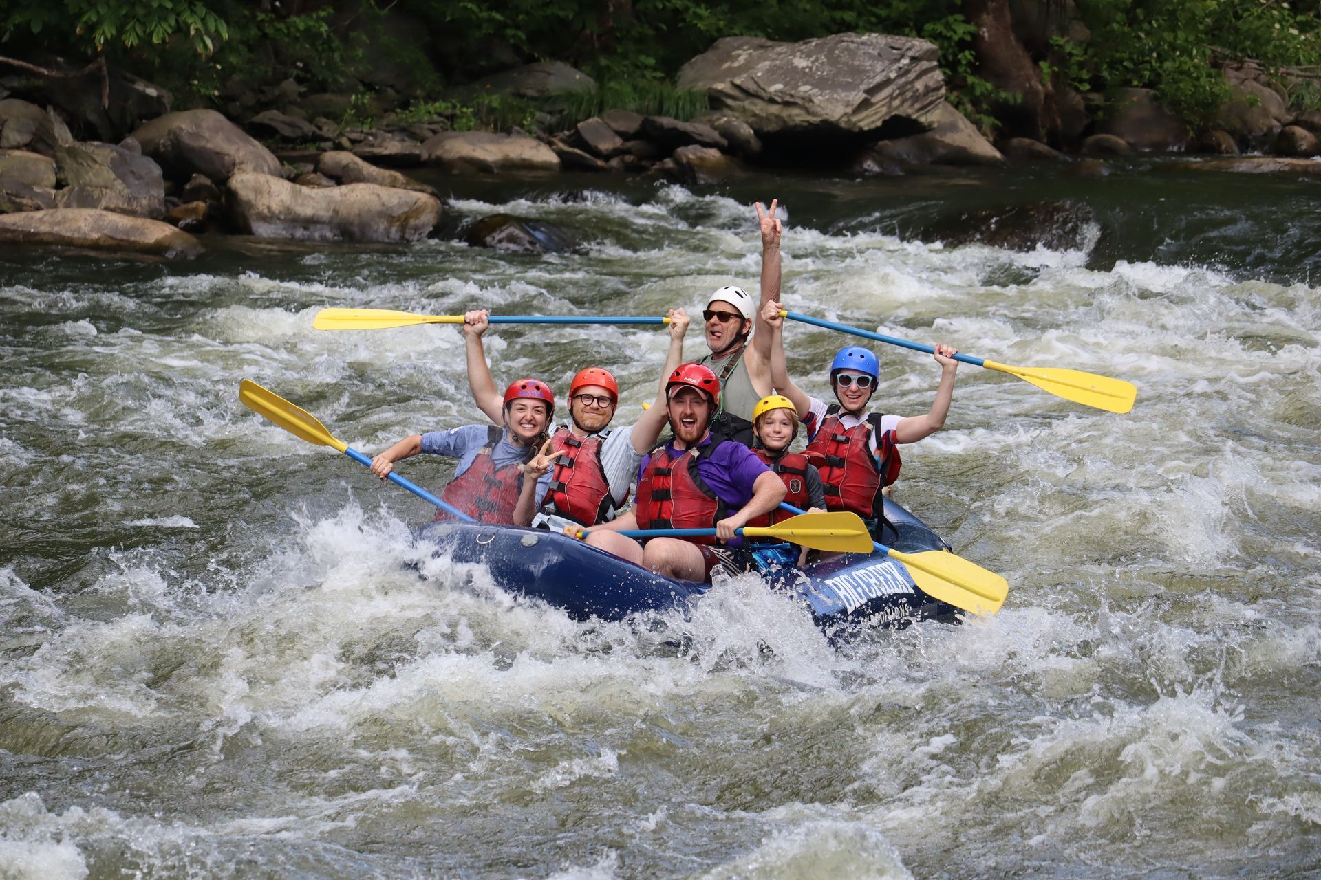 A group in a blue raft navigates river rapids, wearing life vests and helmets while paddling and smiling.