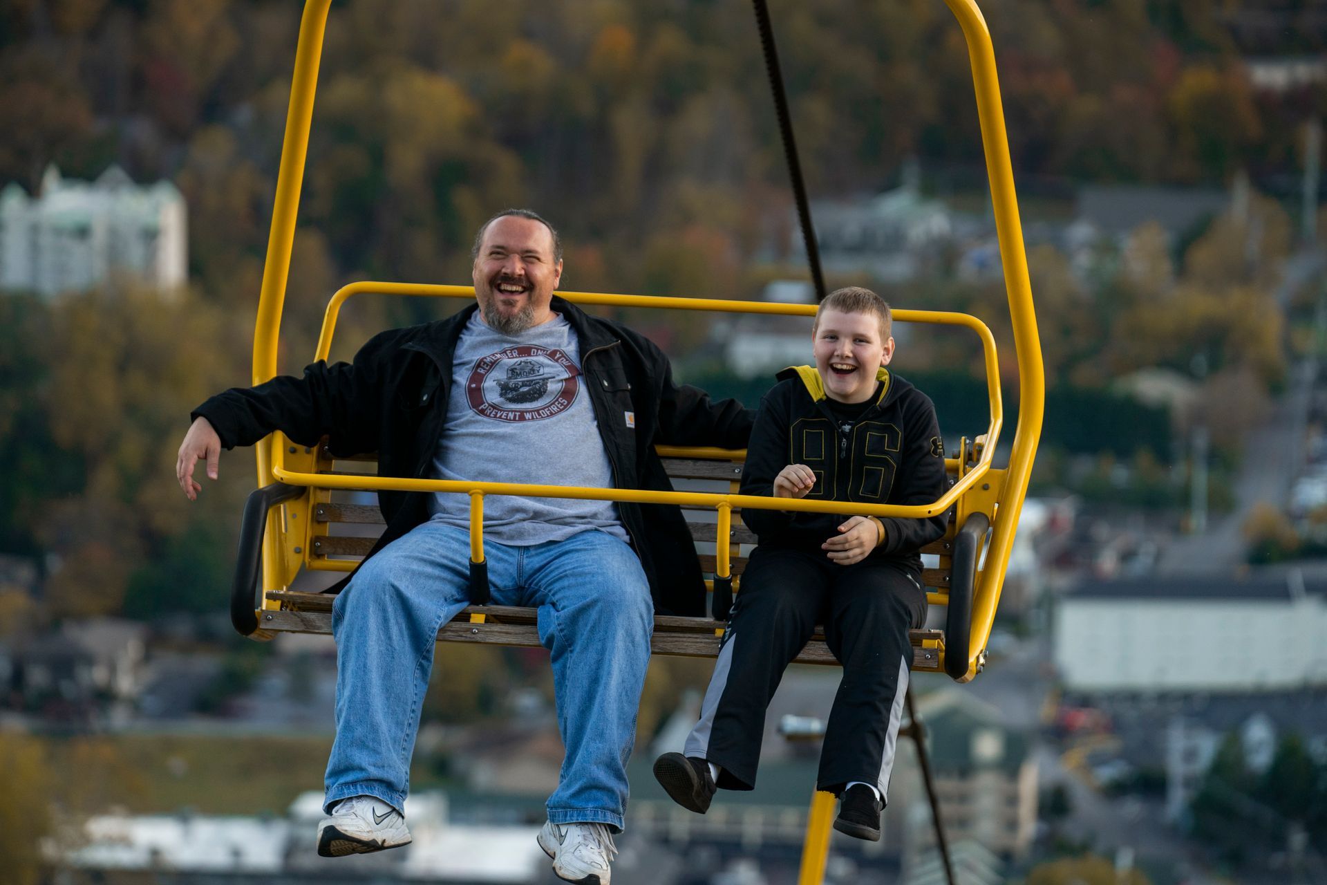 Two people sit side-by-side in a yellow chairlift, laughing with an outdoor town landscape in the background.