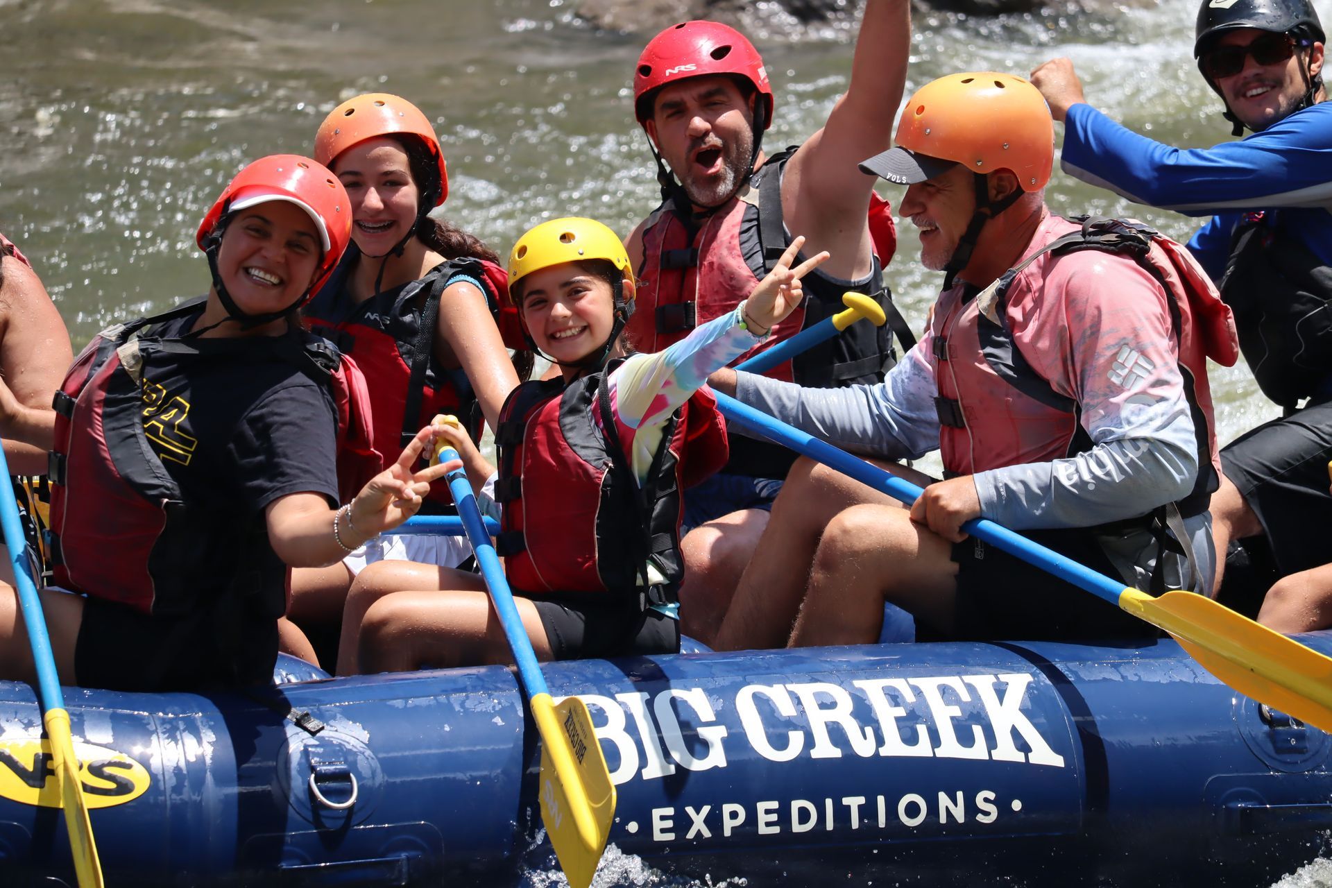 Smiling rafters in helmets on a blue raft labeled “BIG CREEK EXPEDITIONS,” paddling through rapids