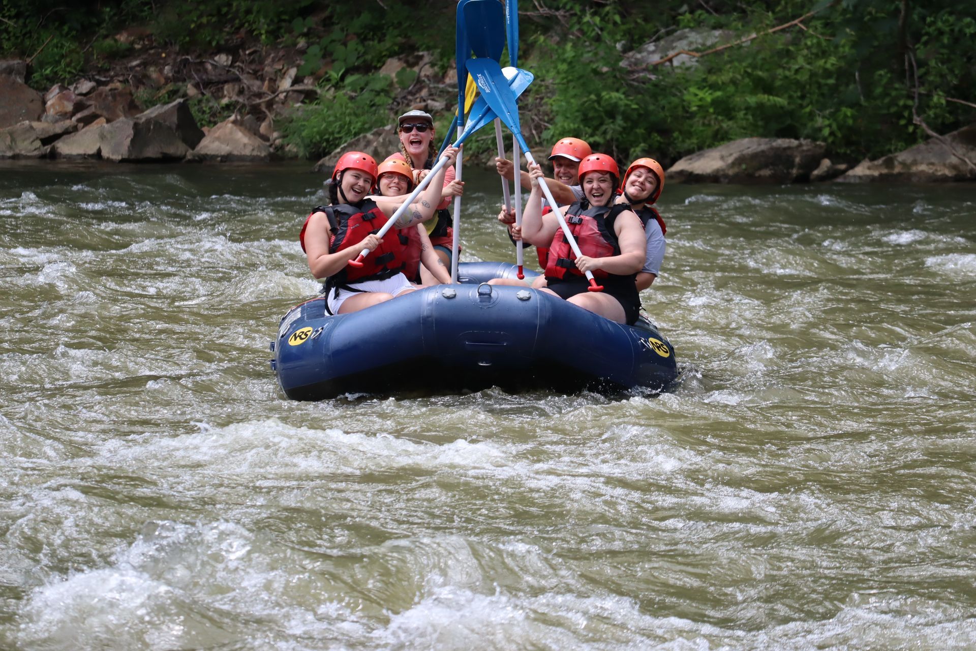 A group of people wearing life jackets and helmets smiles while white-water rafting on a river.