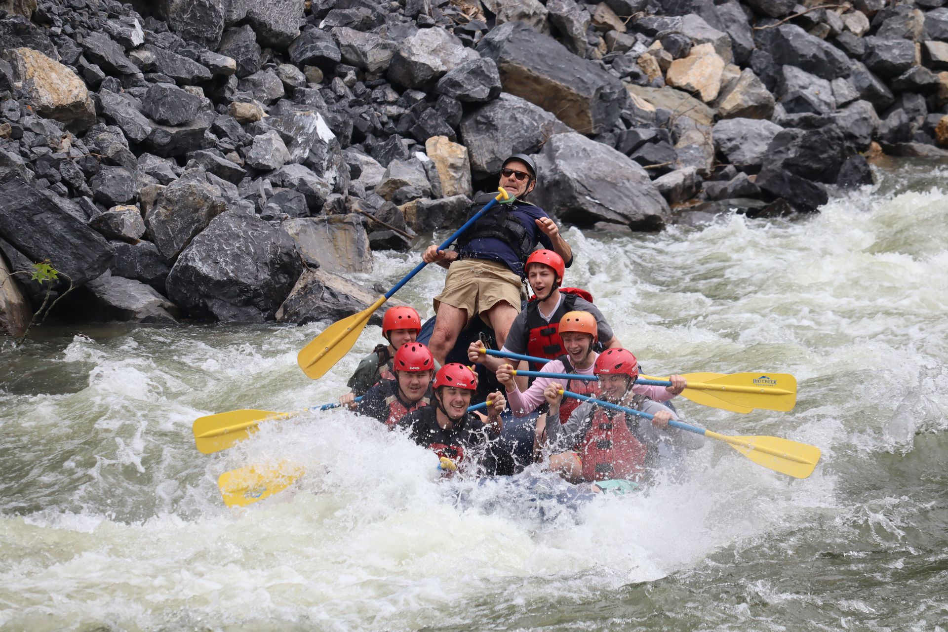 A group of people wearing helmets and life vests whitewater rafting down a rocky river in a yellow raft.