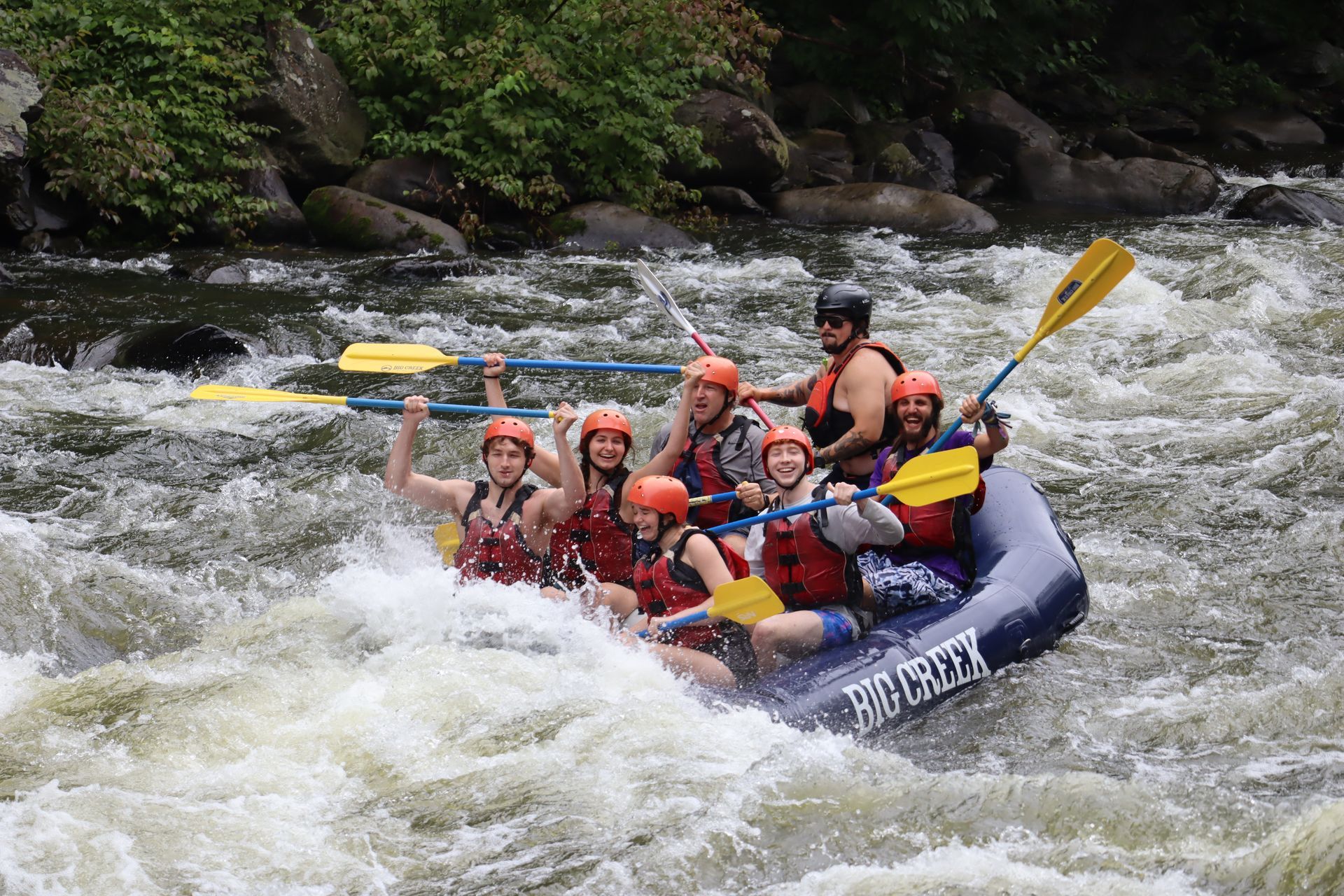A group of people wearing helmets and life vests paddles a raft through white water rapids on a rocky river.