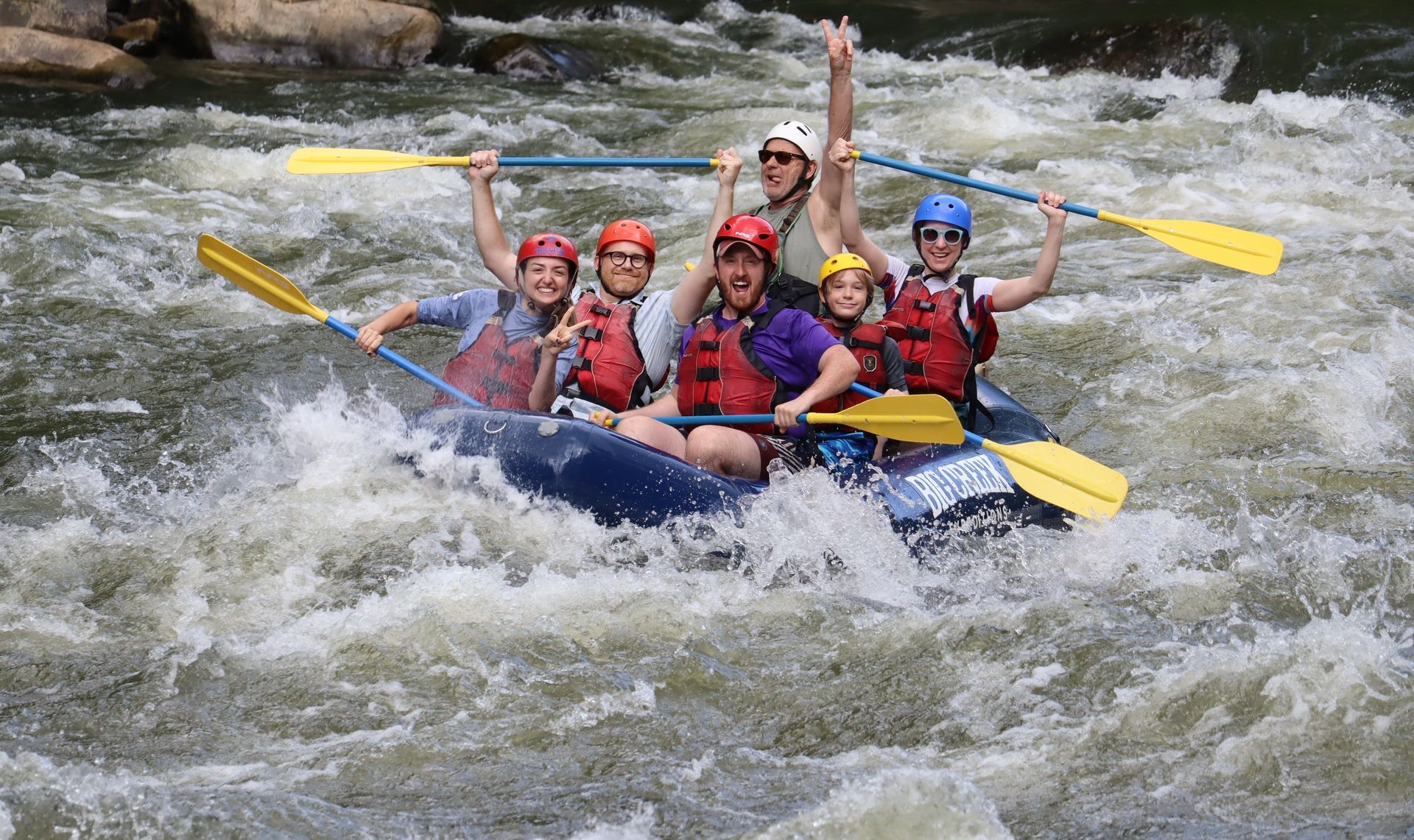 A group of six people in life jackets and helmets whitewater rafting together in a blue raft, smiling and paddling.