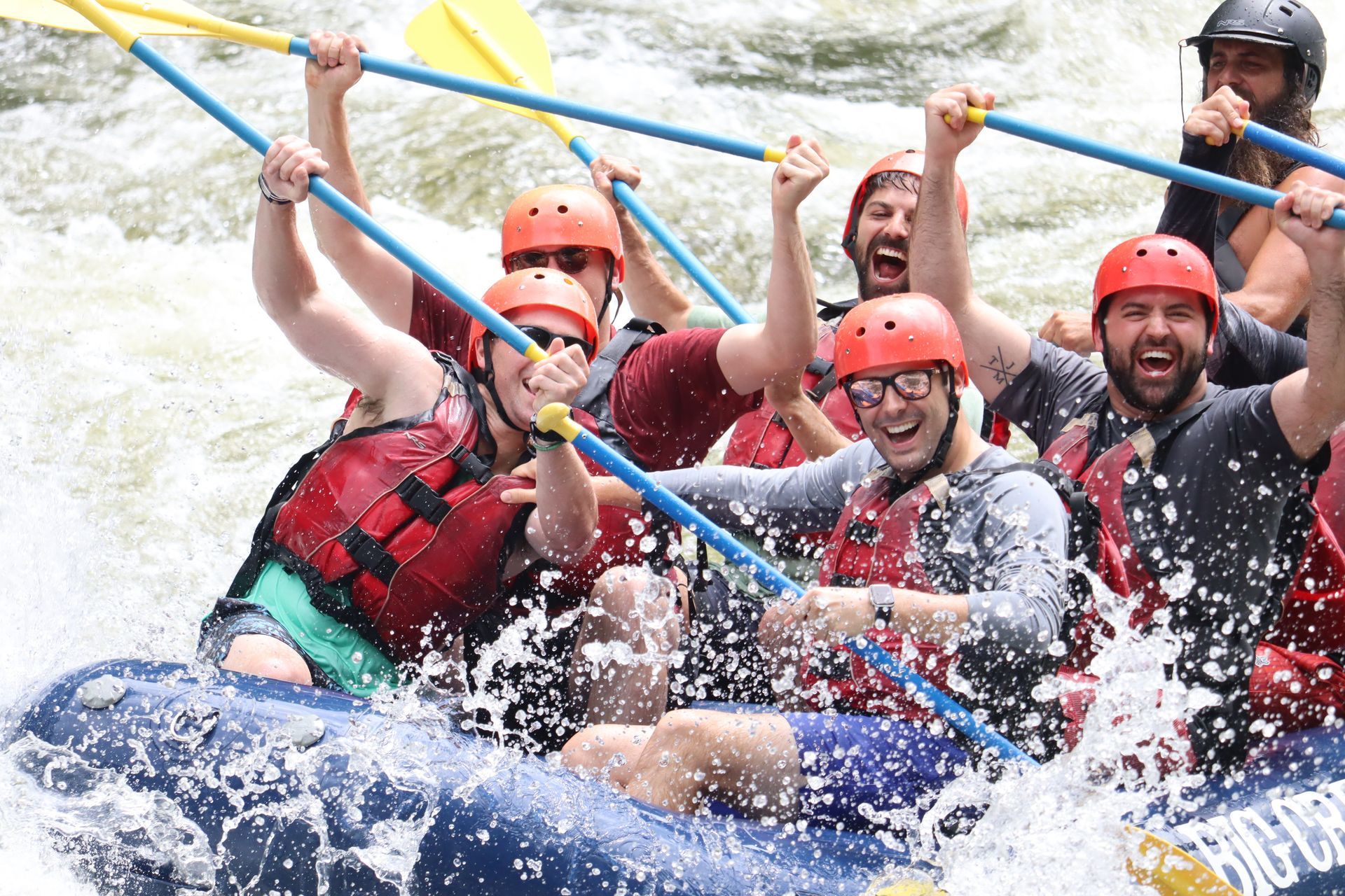 A group of people wearing helmets and life vests cheering while whitewater rafting on a fast-moving river.