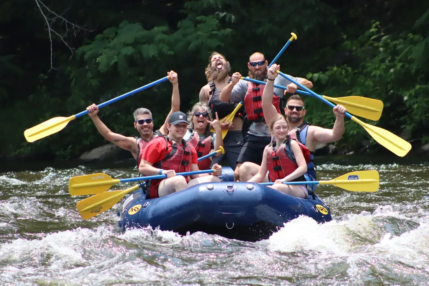 A group of people wearing life jackets cheer while rafting down a river with paddles raised.