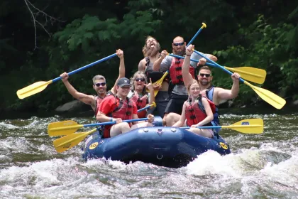 A group of people wearing life jackets cheer while rafting down a river with paddles raised.