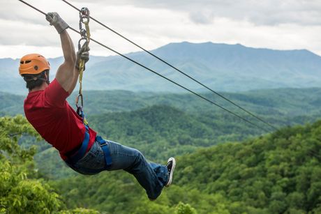 Person zip-lining over a forested mountain landscape, wearing a helmet and red shirt