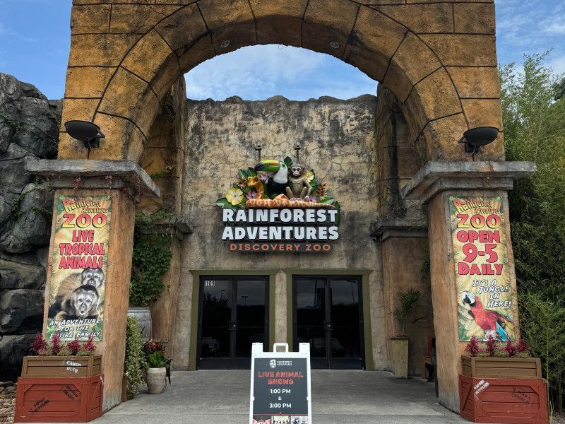 The stone-arch entrance to Rainforest Adventures Discovery Zoo featuring a sign with the zoo's name and hours.