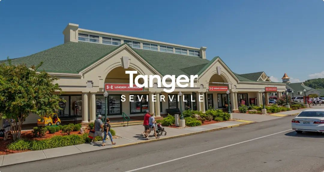 A view of the Tanger Outlets in Sevierville with shoppers walking along the sidewalk in front of stores under a blue sky.