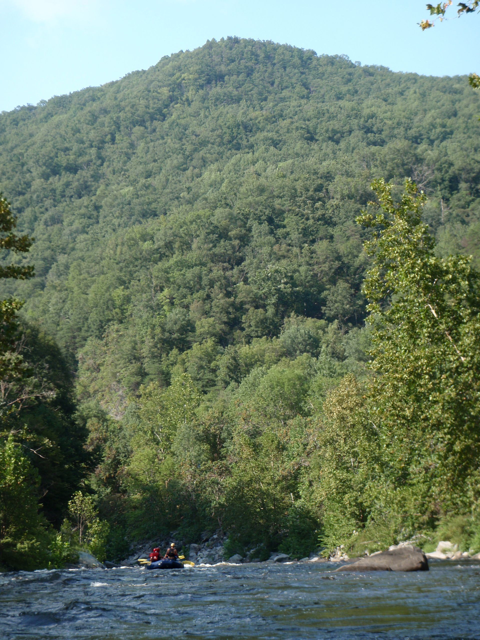 A raft with people floats down a river toward a large, lush green mountain under a clear blue sky.