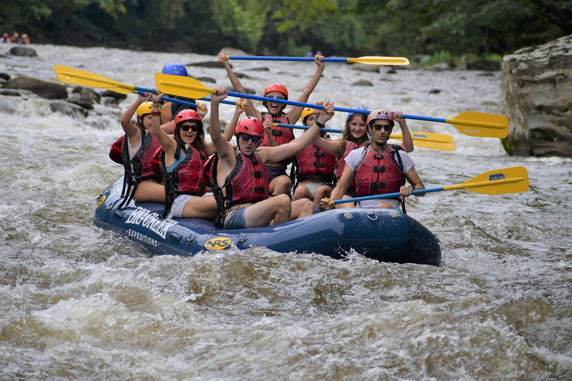 A group of people wearing life jackets and helmets paddle an inflatable raft through rushing river rapids.