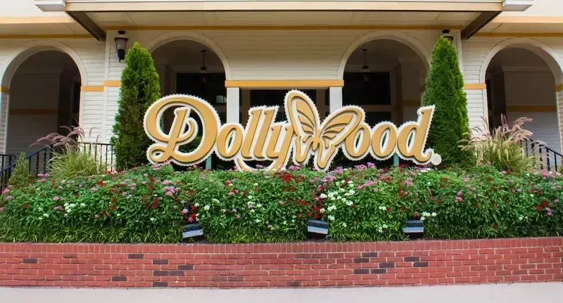 A gold Dollywood sign set in front of flowerbeds and a building with arched entryways.