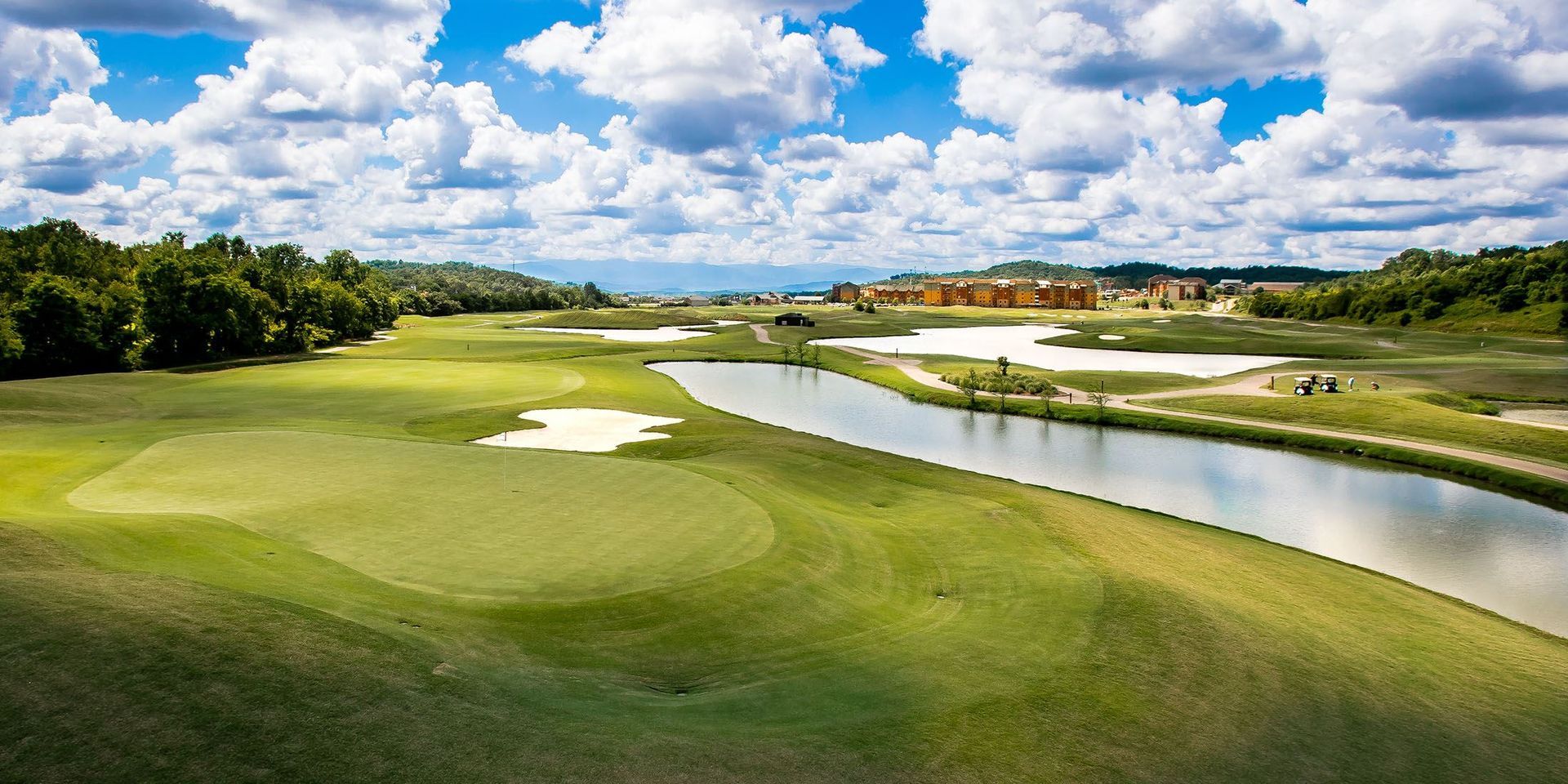 A scenic golf course with a long pond running along the fairway, surrounded by lush grass, sand bunkers, and cloudy skies.