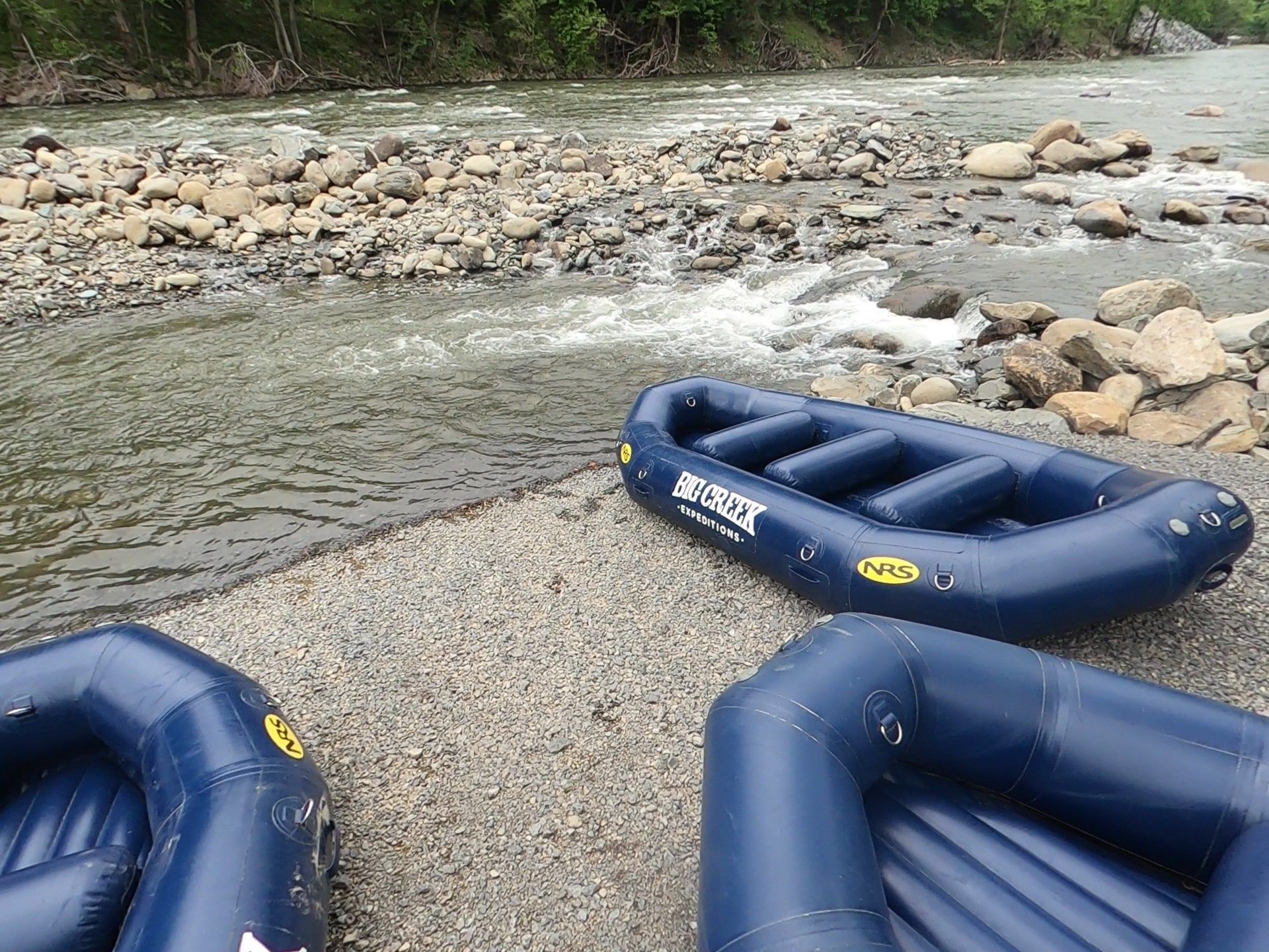 Blue inflatable rafts on a rocky riverbank beside shallow flowing water and trees