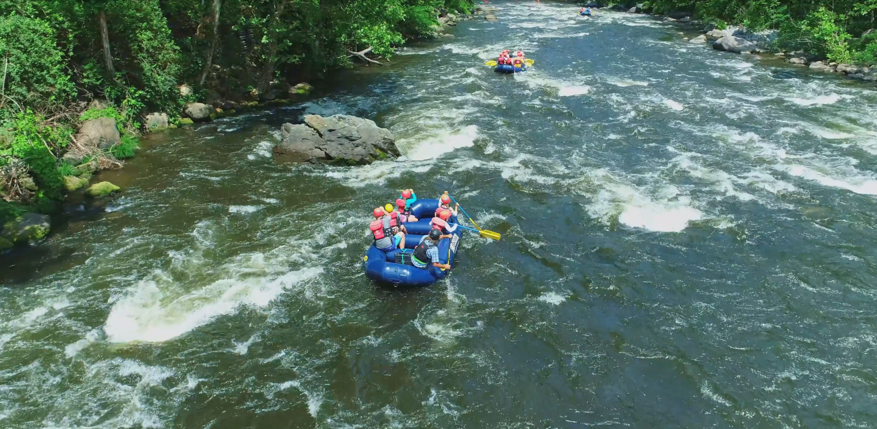An aerial view of two blue rafts with people in life jackets navigating a river with light rapids through a wooded area.