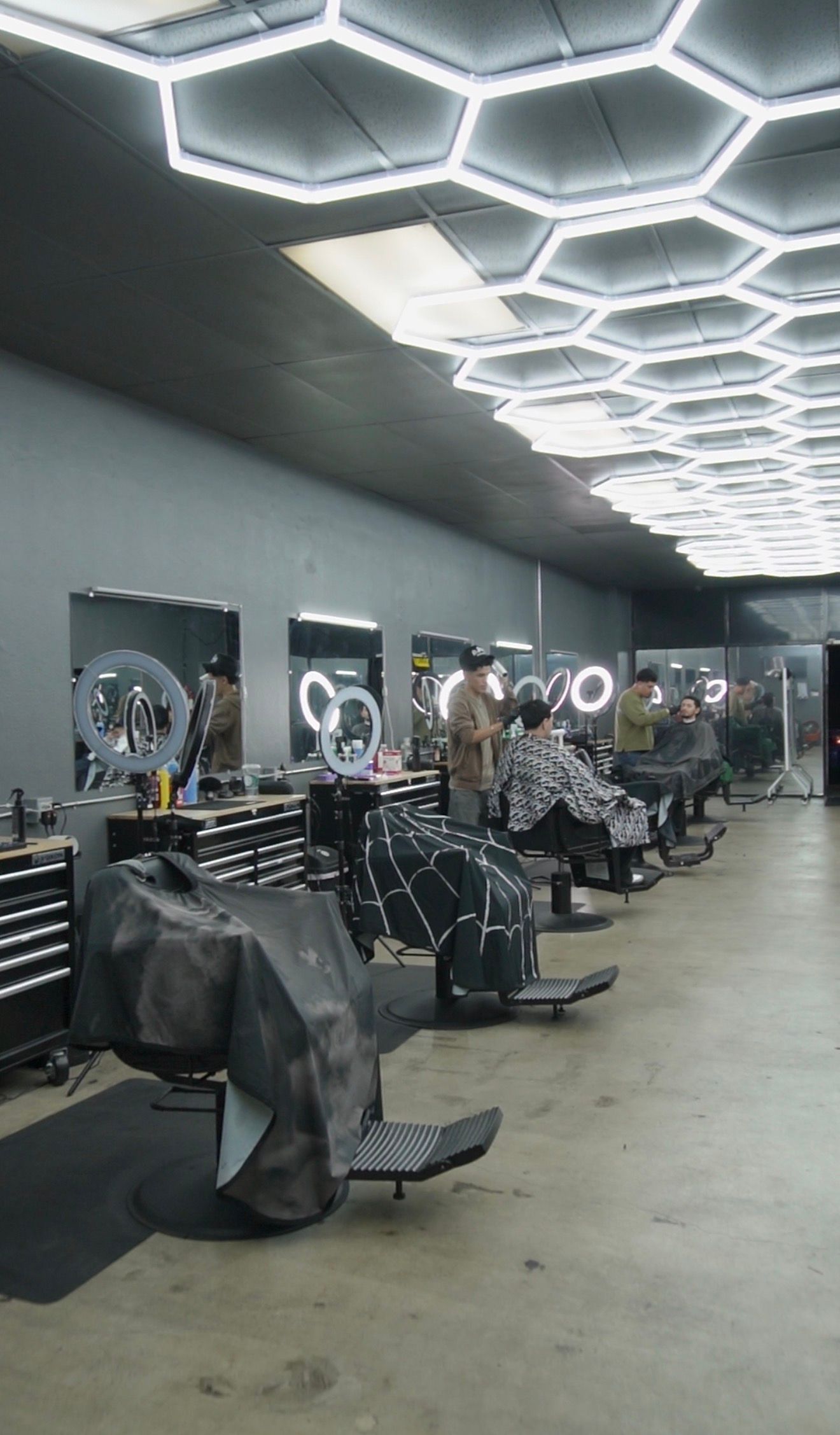 A barber shop interior with several stations. Black chairs, mirrors, and hexagon-shaped lights illuminate the gray space.