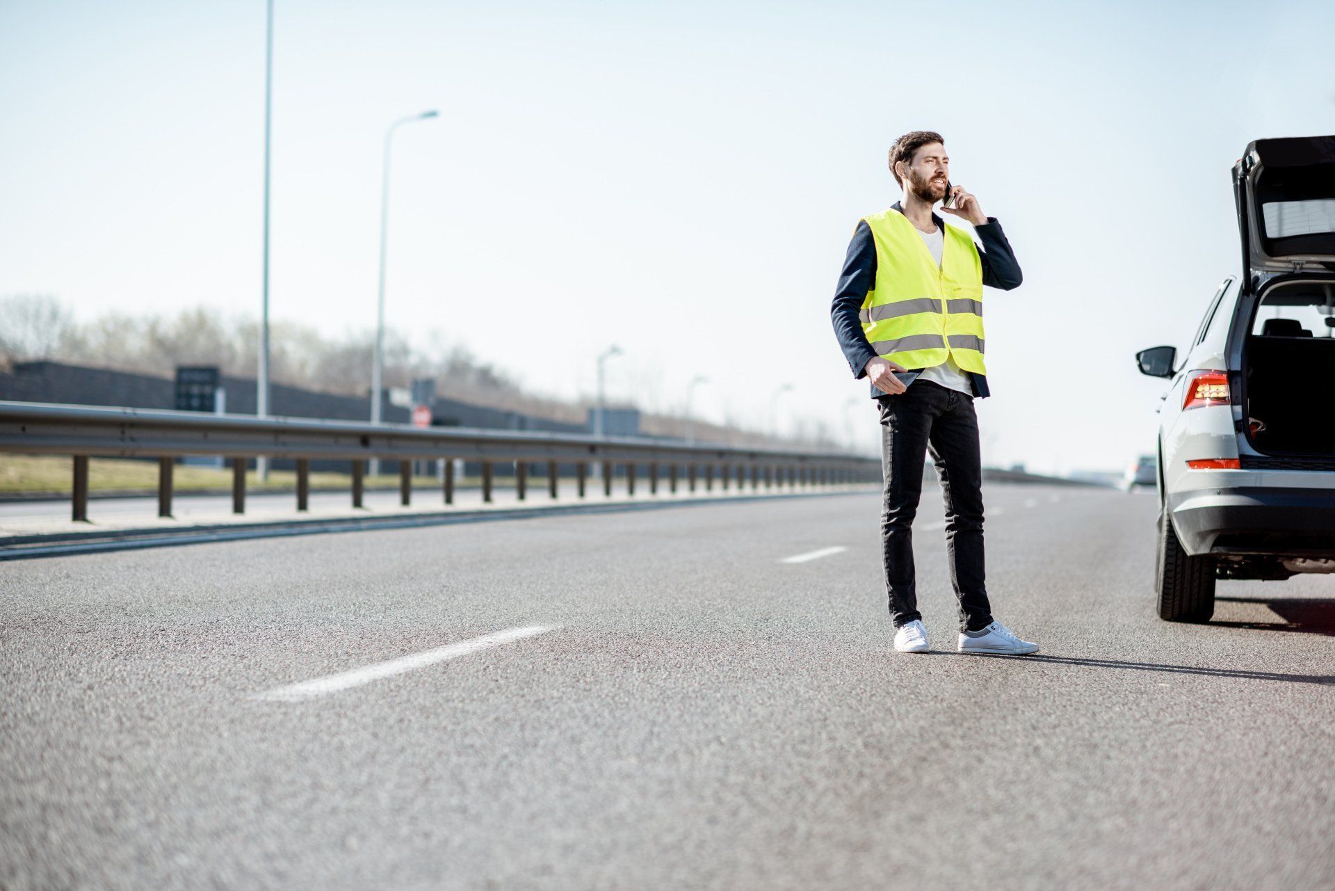 Uomo con giubbotto catarifrangente in piedi accanto a un'auto in panne sulla corsia di emergenza dell'autostrada, intento a usare il telefono.