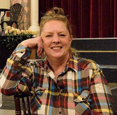 A woman in a plaid shirt is sitting at a table with her hand on her chin.