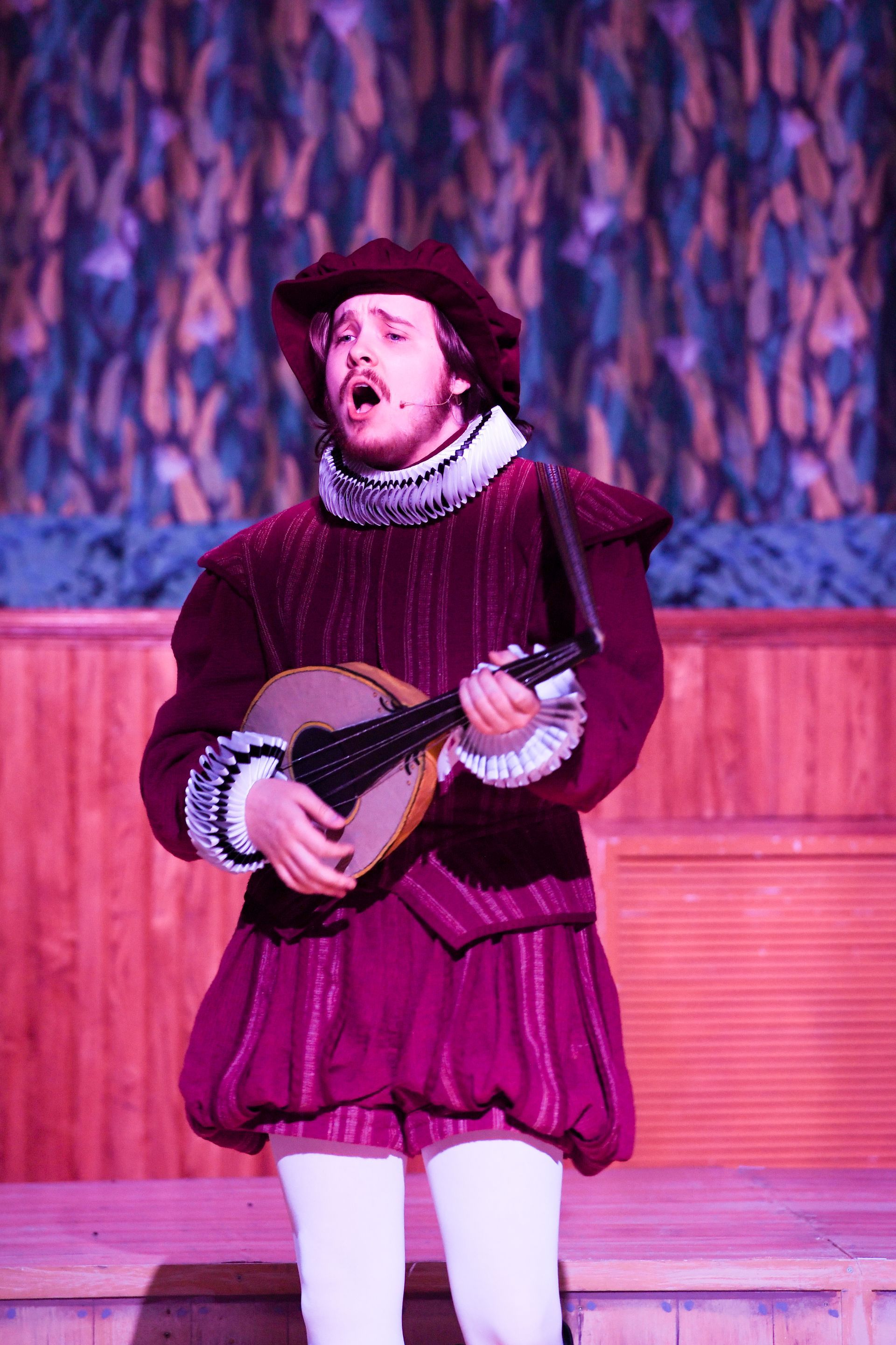A man in a purple dress is holding a mandolin and singing on a stage.