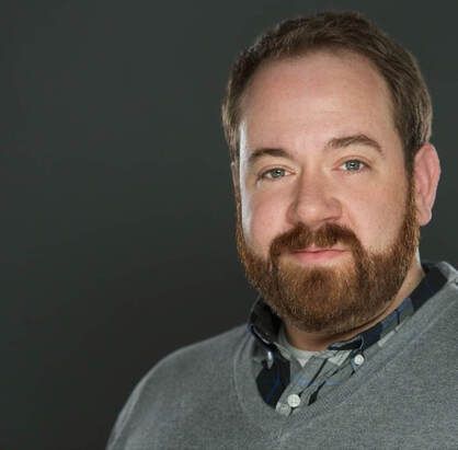 A man with a beard is wearing a grey sweater and a plaid shirt.