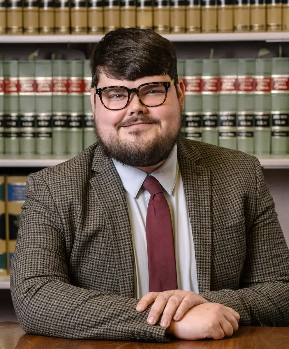 A man in a suit and tie is sitting at a table in front of a bookshelf.