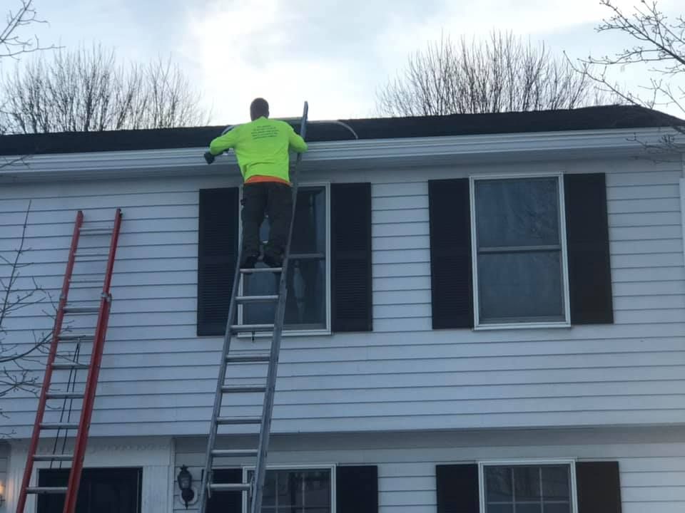 Person on a ladder, cleaning gutters on a two-story white house with black shutters.