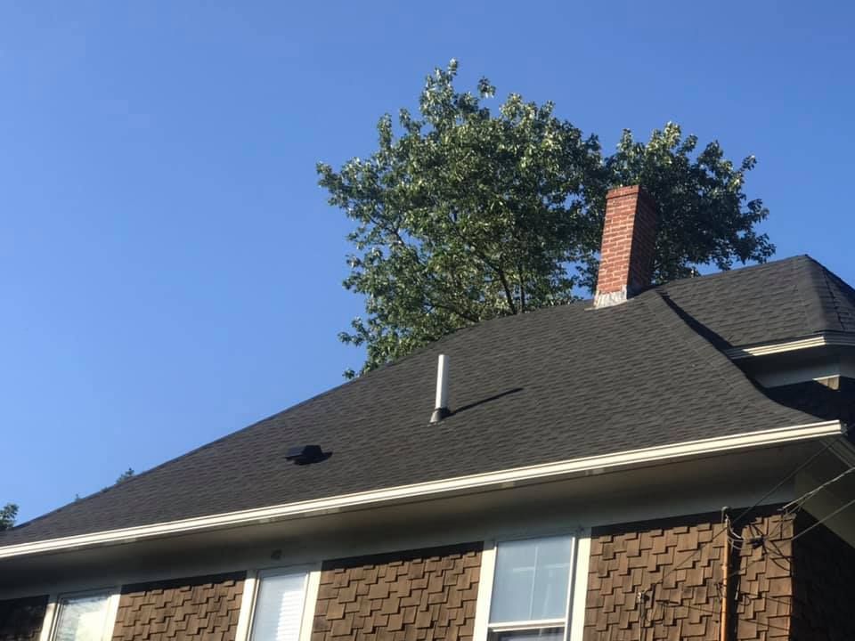 A house with a black shingled roof, a chimney, and a tree against a blue sky.