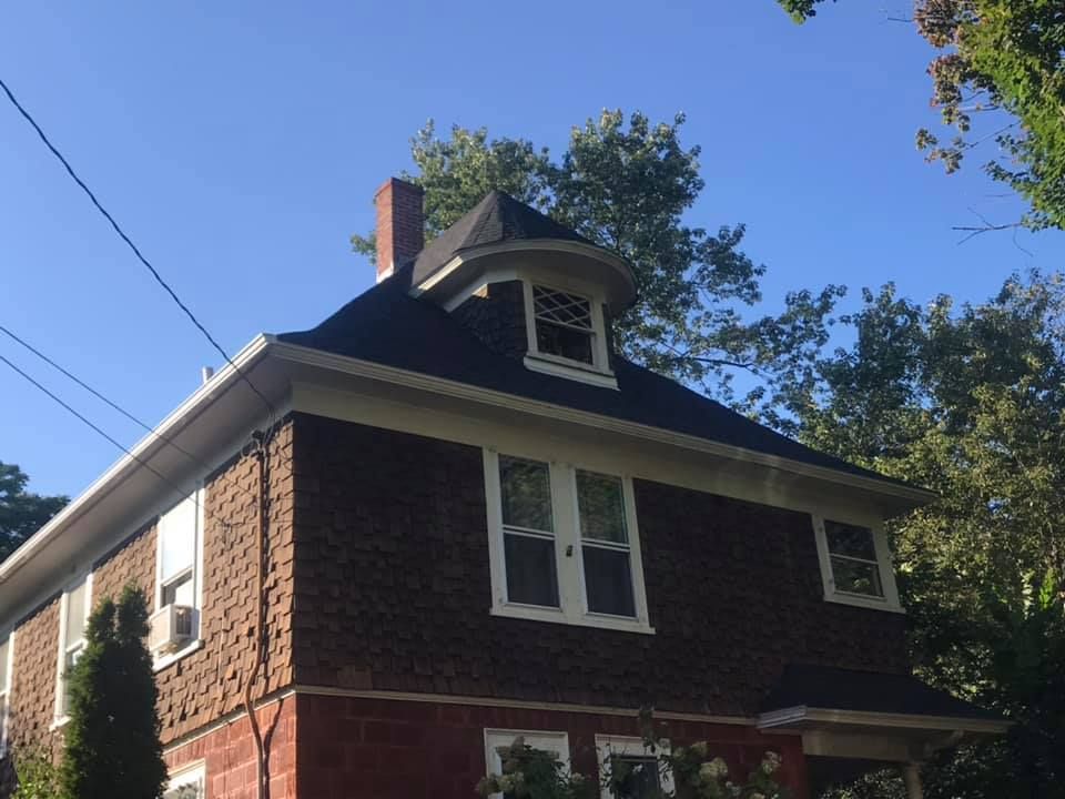 Two-story house with brown siding, a dark roof, and a turret-like dormer with a small window under a bright blue sky.